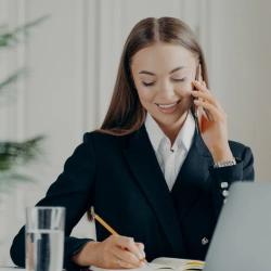 Młoda kobieta w czarnym żakiecie i białej koszuli rozmawia przez telefon komórkowy przy uchu i robi notatki ołówkiem w notesie przy biurku.